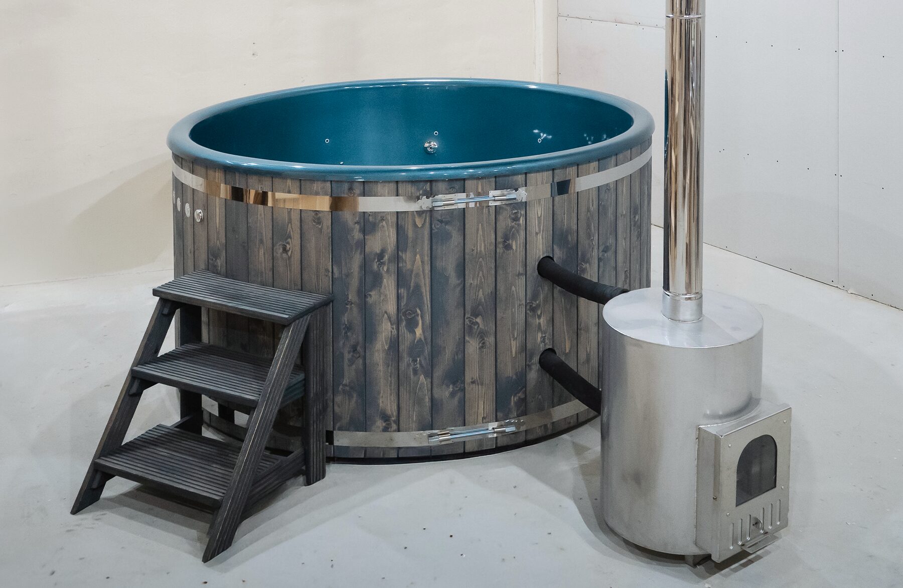 Makoya Spa hot tub DIANA is placed in the corner of a room, featuring a wooden cladding, wooden entry steps, an isolating cover and a external metal stove with a tall chimney