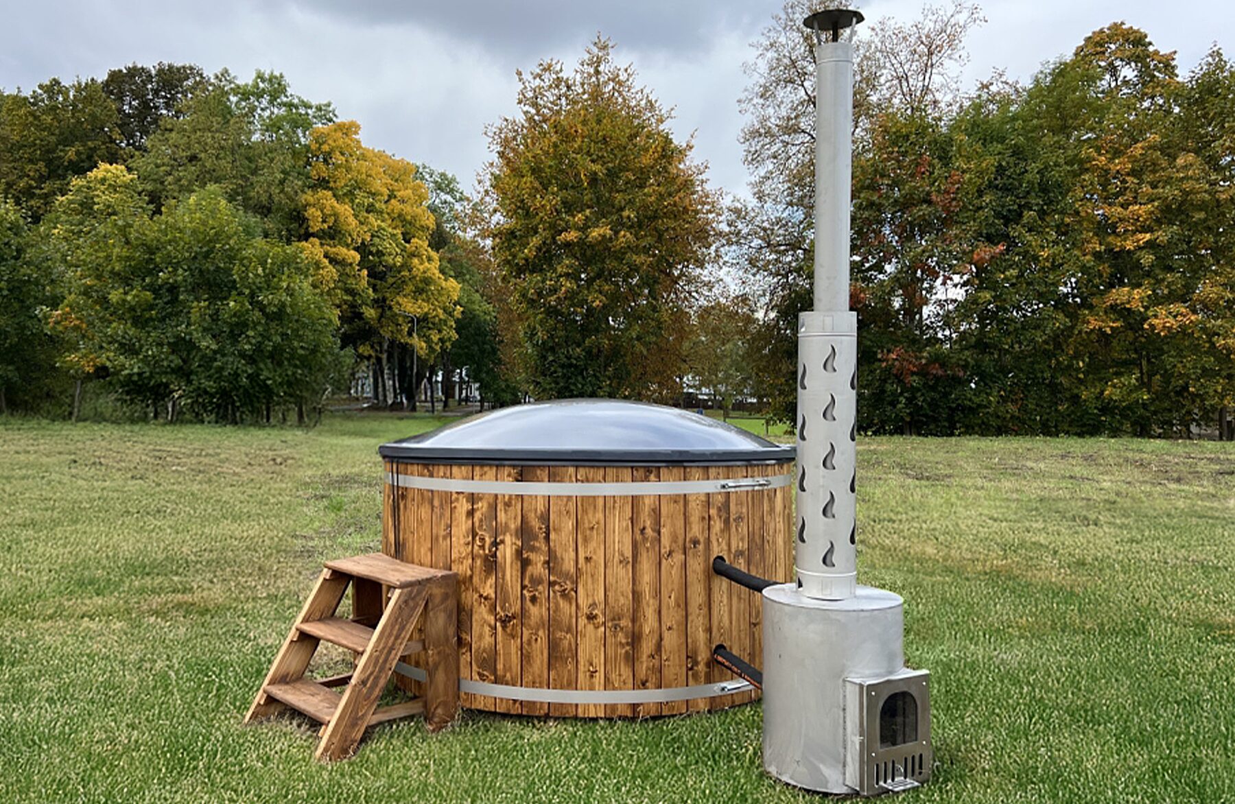 Makoya Spa hot tub DIANA is placed on a green meadow, featuring a wooden cladding, wooden entry steps, an isolating cover and a external metal stove with a tall chimney, sourrounded by a natural landscape