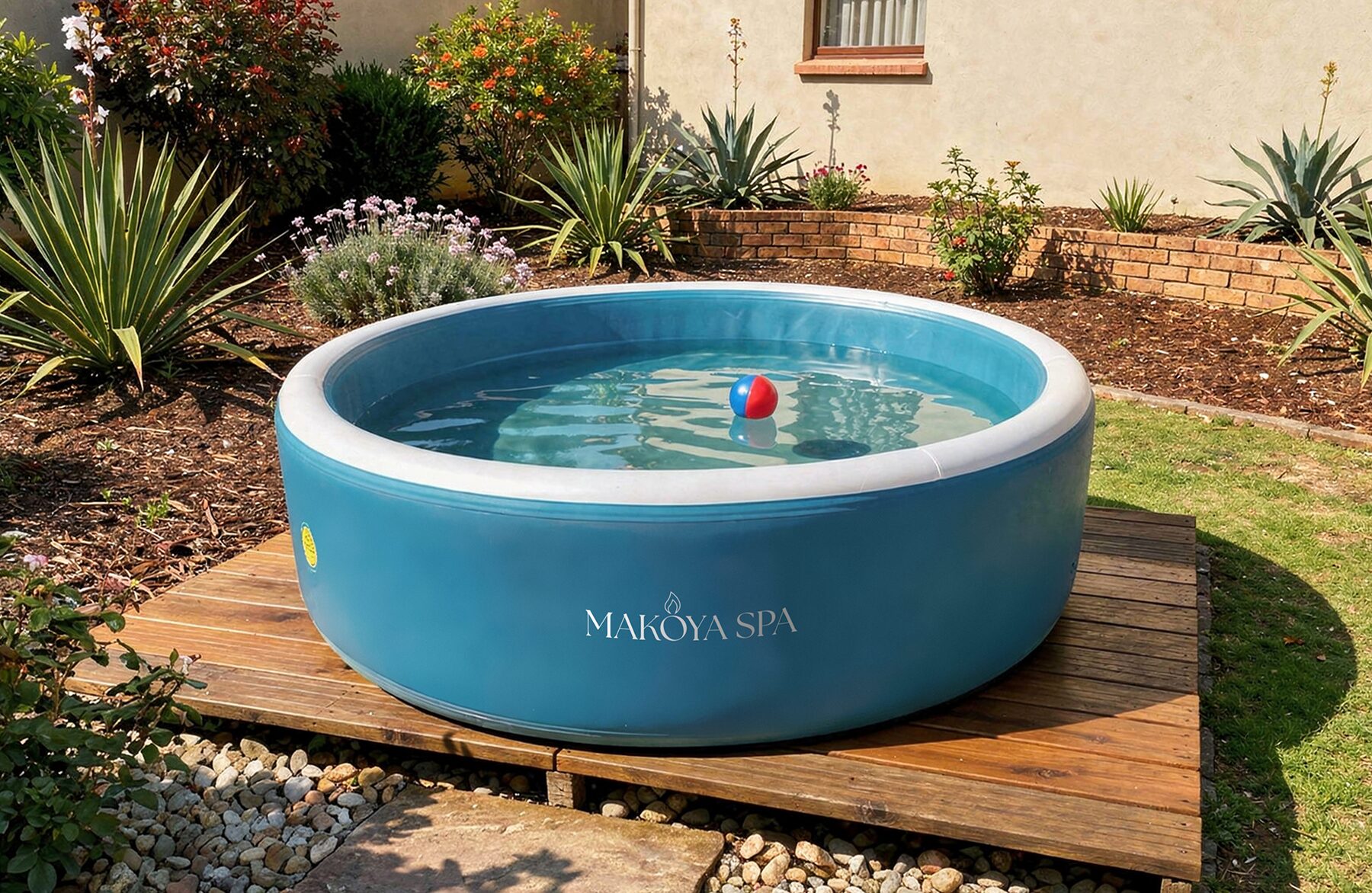 Exterior view of a round inflatable pool by Makoya Spa with a blue outer wall and clear water, placed on a wooden deck in a well-maintained garden with green lawn, flowering plants, and a house in the background.