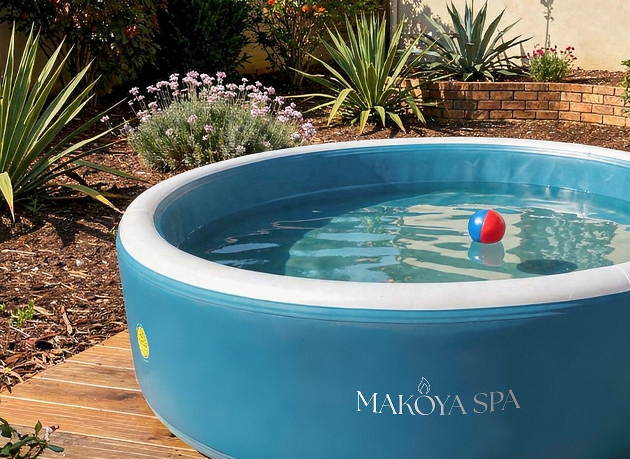 Exterior view of a round pool by Makoya Spa with a blue outer wall and clear water, placed on a wooden deck in a well-maintained garden with green lawn, flowering plants, and a house in the background.