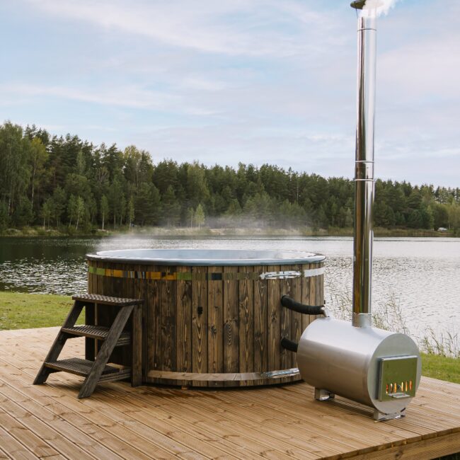 Wooden hot tub LUNA by Makoya Spa on a wooden deck, with external metal stove and chimney, wooden steps and green natural landscape in the background.