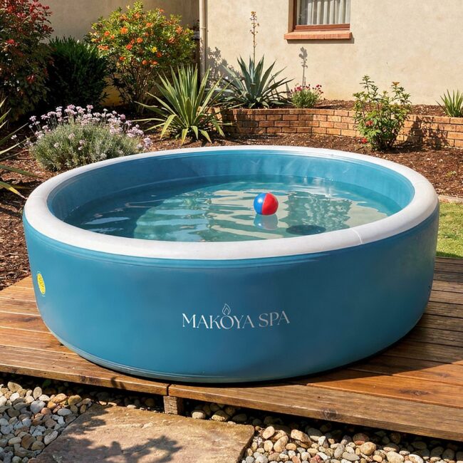 Exterior view of a round inflatable pool by Makoya Spa with a blue outer wall and clear water, placed on a wooden deck in a well-maintained garden with green lawn, flowering plants, and a house in the background.