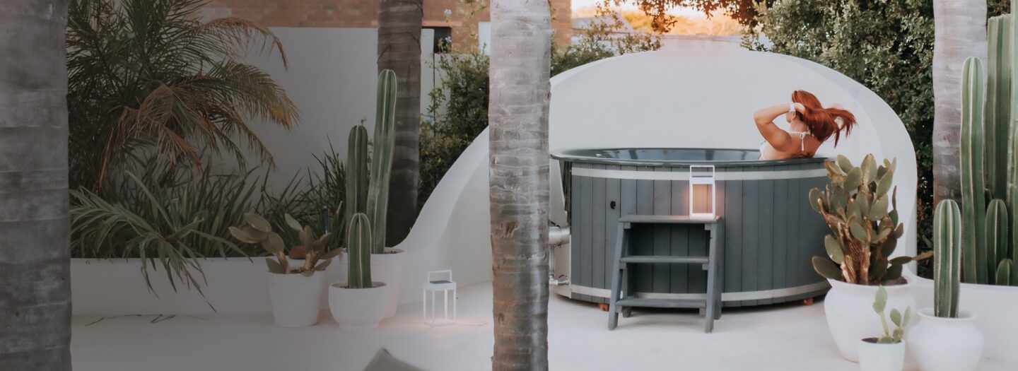 A person relaxing in a wood-fired hot tub by Makoya Spa, in a garden with palm trees and cacti.